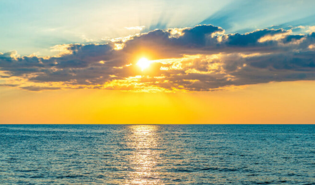 The image shows a serene sunset over the ocean with vibrant blue and orange skies, clouds around the sun, and light reflecting on water.