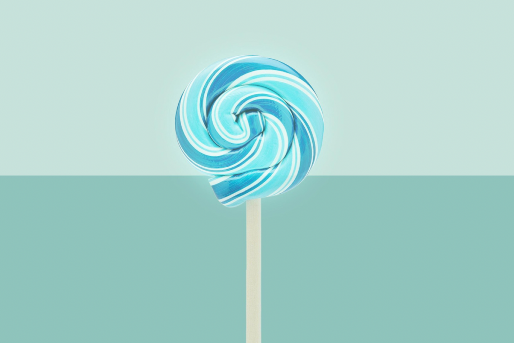 A blue and white swirled lollipop stands centered on a wooden stick against a two-tone light teal background, creating a minimalist and playful visual.
