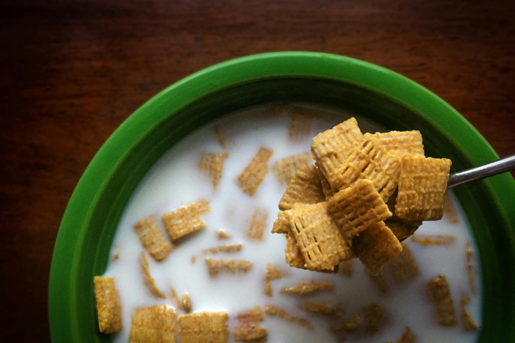 A bowl of square-shaped cereal with milk, atop a wooden surface. A spoon holds some cereal pieces, and the bowl rim is green.